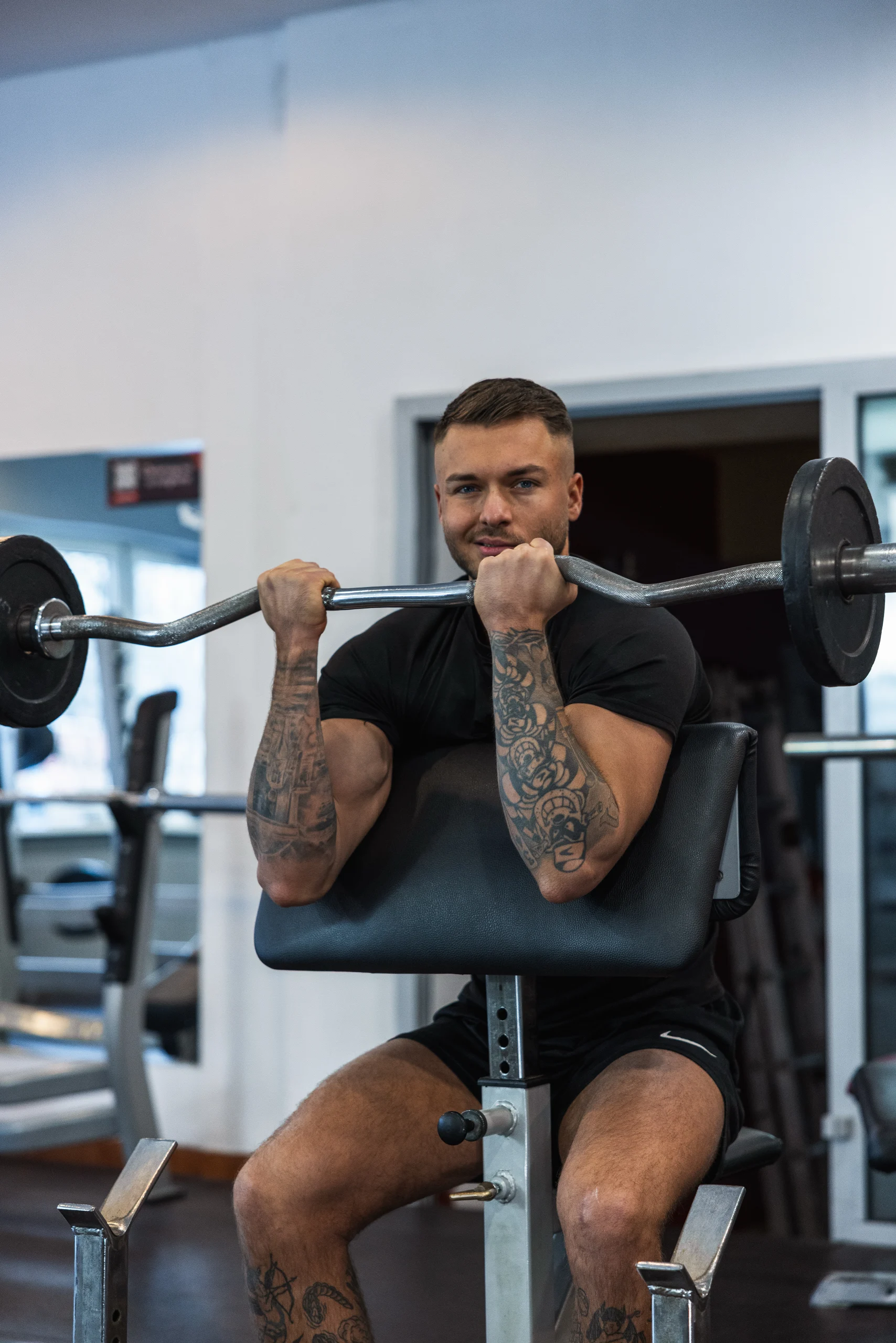 Trener Michał wyciskający sztangę na biceps i oparty o modlitewnik, w tle siłownia z lustrami.