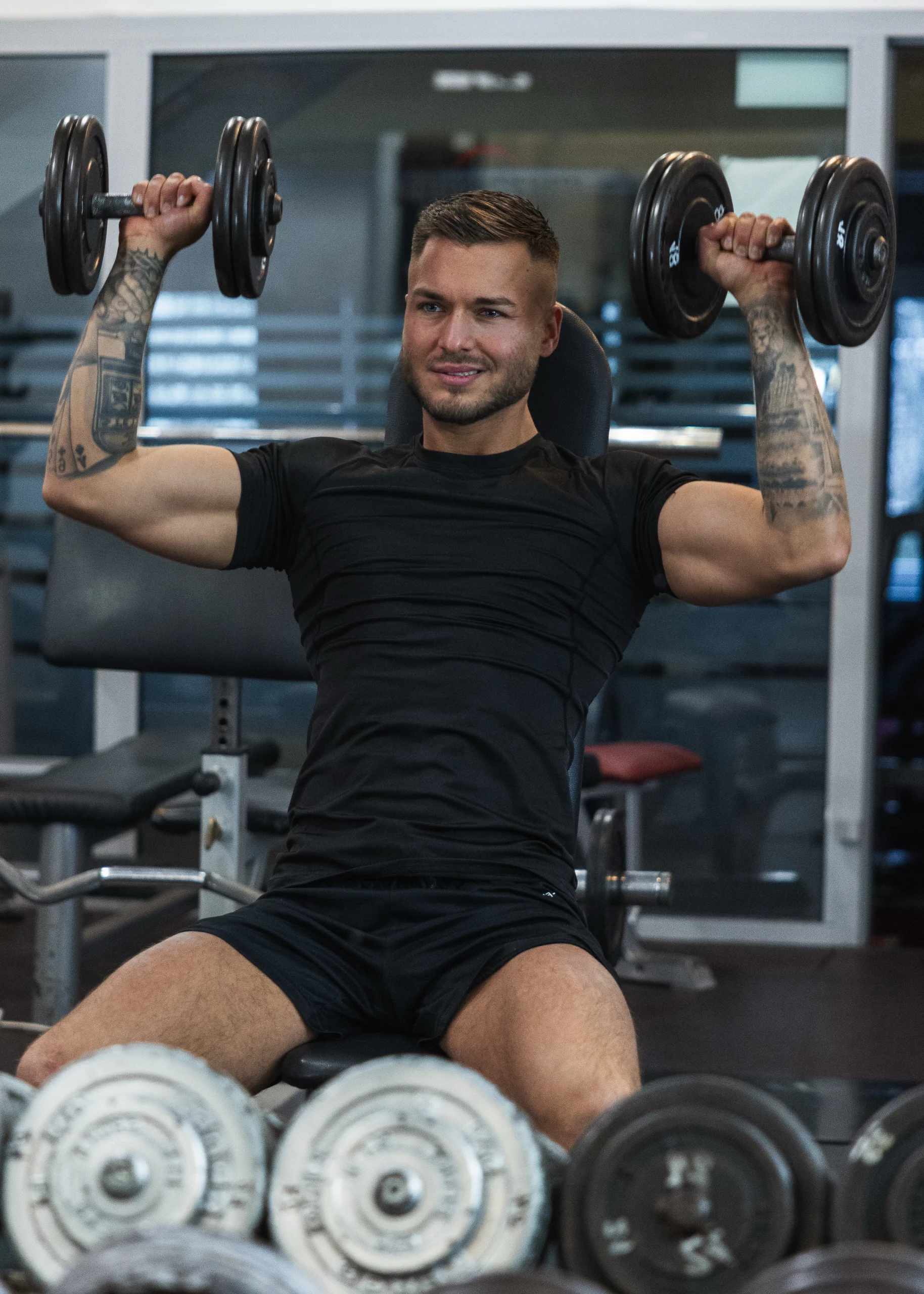 Trener Michał trzymający wyciskający hantle w górę na tle siłowni.